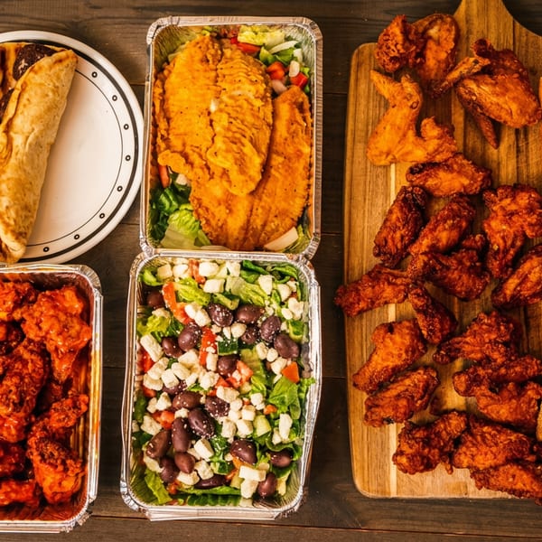 Fried chicken wings, fish fillets, salads, and submarine sandwiches on a rustic wooden table.