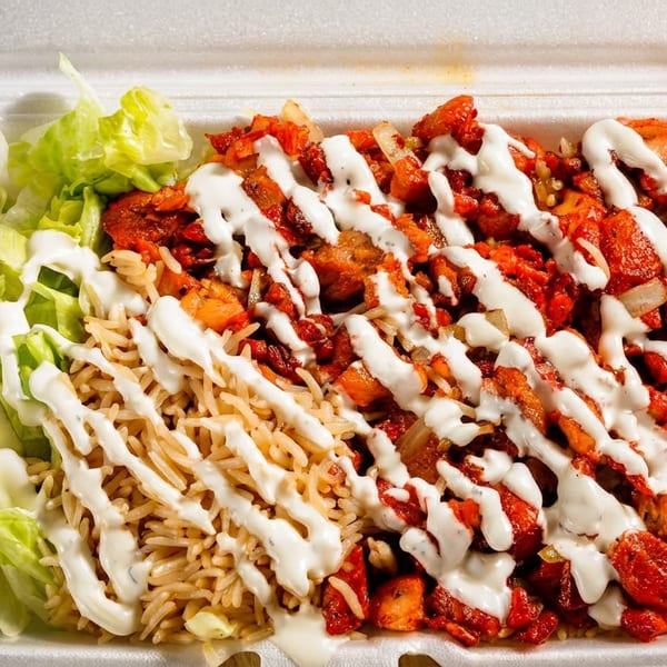 Spicy red chicken and rice platter with lettuce, white sauce, and side sauces in a takeout container.